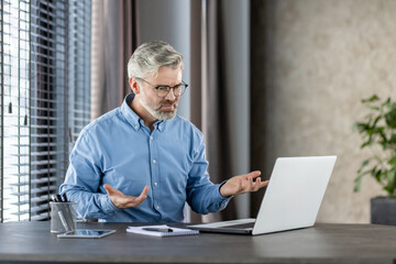 Senior businessman expressing frustration during a video call on a laptop in a modern office with large windows and a plant.