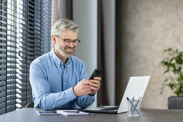 Smiling mature businessman using smartphone while sitting at office desk with laptop and notebook....