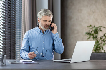 Mature businessman in blue shirt talking on phone while working on laptop at office desk....