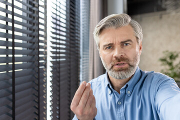 Mature man with gray hair and beard looking serious and gesturing near a window with blinds....