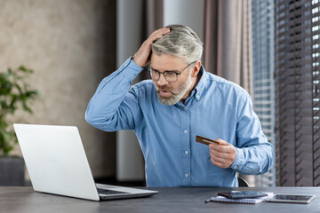 Older man holding a credit card and looking shocked at a laptop screen, sitting indoors. Concept of...