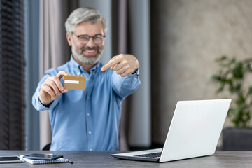 Smiling man presenting credit card next to a laptop in a home office setting. Concept of online...