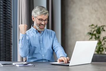 Happy man in blue shirt celebrating successful work outcome while using laptop in home office...