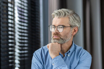 Mature man with glasses looking thoughtful while standing by a window in a bright room. Reflective and contemplative expression