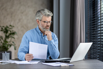 Senior professional man working on a laptop while holding documents, depicting thoughtful...