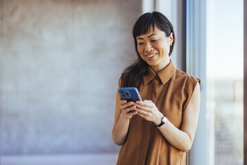 A professional Asian businesswoman smiles as she reads her smartphone, wearing casual office attire, reflecting ease and productivity in her corporate environment.