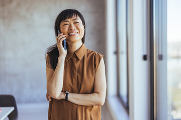 Joyful Asian female professional in smart casual wear engages in conversation on her smartphone, standing by the window in a well-lit contemporary workspace.