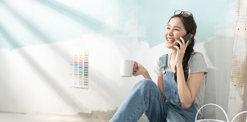 Smiling asian woman using phone after painting in room at new home.Asian young woman resting with...