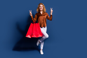 Full size photo of positive girl wear silk blouse jeans show v-sign symnol hold shopping bags isolated on dark blue color background