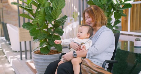 Fototapeta premium A loving mother lifts her smiling baby for a kiss in cozy indoor with lush green plants. This heartwarming moment captures the essence of maternal affection joy in a beautiful natural environment