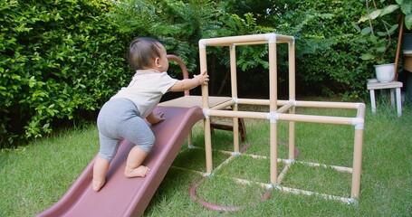 A happy Asian baby toddler child is playing on a slide in a backyard. The baby is crawling up the...