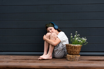 Cute pre-teen boy in headphones listening to music sitting on a wooden terrace hugging his knees