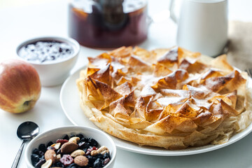 Sweet, apple pie made of phyllo dough on a white plate, on the table with a napkin, next to nuts, jam and a cup of tea.