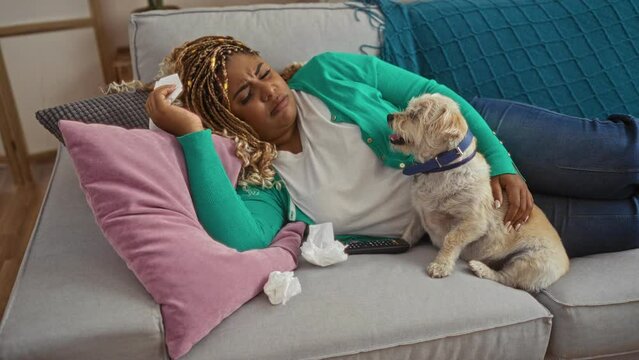 A young african-american woman with braids, lying on a couch with a dog, is watching a sad movie, wiping tears with a tissue in a cozy living room at home.