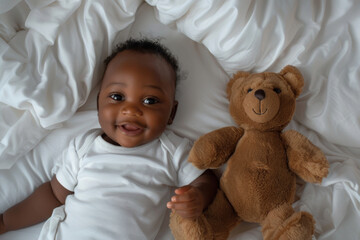 Portrait of cute smiling African American baby and toy teddy bear lying on white bed at home. Top view