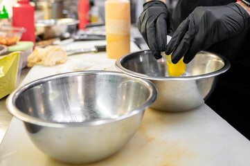 A person in a kitchen, wearing black gloves, cracking an egg into a bowl amidst food items