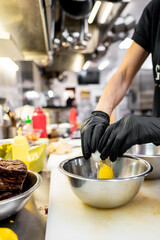 A person in a kitchen, wearing black gloves, cracking an egg into a bowl amidst food items