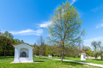 Fototapeta premium Small chapels at Skalka Calvary stand amidst vibrant green trees under a clear blue sky, signaling the arrival of spring. Mnisek pod Brdy, Czechia