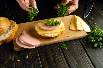 Chef hands adding parsley to a sandwich with sausage and cheese on the kitchen table. Concept of preparing a quick meal in a bar kitchen.