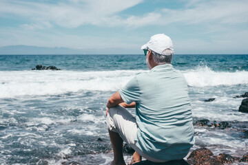 Rear view of carefree senior relaxed man barefoot wearing  cap sitting on a rocky beach admiring the sea waves crashing on the beach. Travel vacation leisure activity concept