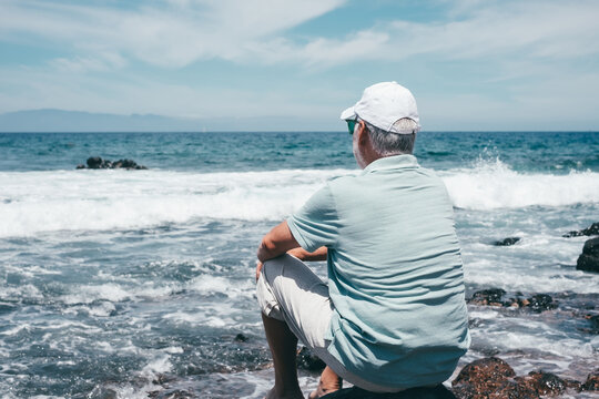 Rear view of carefree senior relaxed man barefoot wearing cap sitting on a rocky beach admiring the sea waves crashing on the beach. Travel vacation leisure activity concept - Powered by Adobe