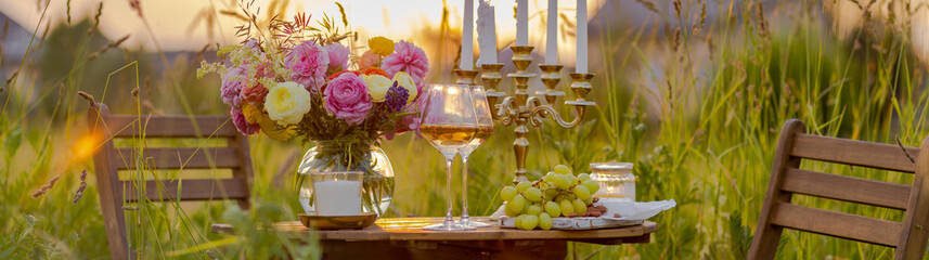 Beautiful romantic outdoor wedding decor in a field. Table decorated with burning candles, flowers. Wineglasses with white wine. Sunset, summer, golden hour. Perfect surprise date for couple. Banner