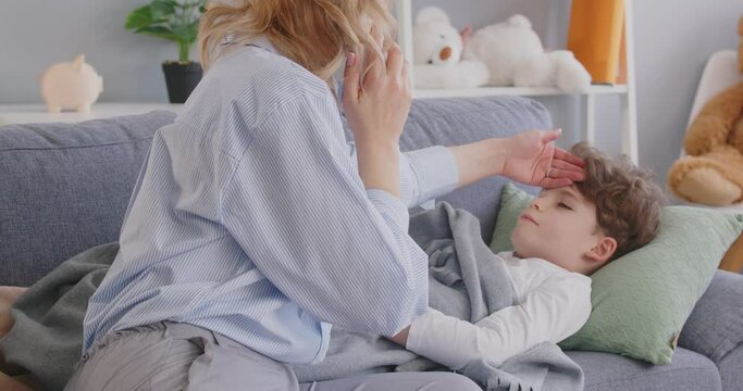Caring mother calls family doctor or pediatrician when her child falls ill. Worried woman sitting by sick son, feeling his forehead, touching skin on his face, calling doctor and telling about fever