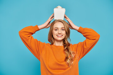 A beautiful woman in an orange sweater joyfully holds box of Asian food over her head.