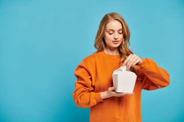 A beautiful blonde woman in an orange sweater holds a white food box.