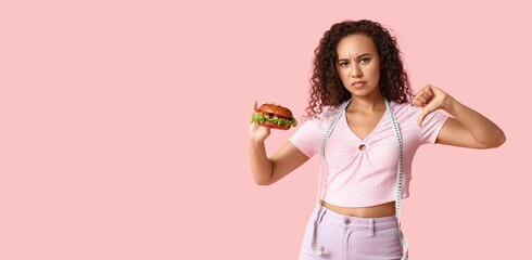 Sad African-American woman with measuring tape and burger showing thumb-down on pink background...
