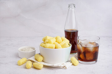 Bowl with tasty salty corn sticks, glass of cola and sauce on light background