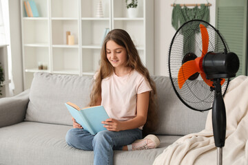Teenage girl sitting on sofa near electric fan and reading book in living room