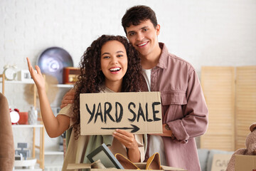 Young couple holding cardboard with text YARD SALE in room of unwanted stuff