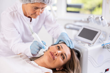 Fototapeta premium Female cosmetologist doing anti-aging face hardware therapy procedure for young female client in clinic of aesthetic medicine