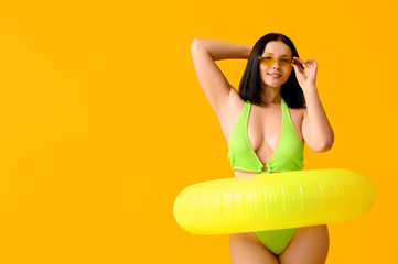 Beautiful young woman in stylish green swimsuit and sunglasses with inflatable ring on yellow...