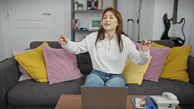 Young woman enjoying music while dancing indoors, surrounded by colorful pillows in a cozy living room.