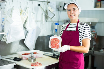 Smiling young female butcher demonstrating freshly formed ground beef hamburger patties in plastic...
