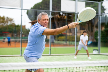 Emotional mature male playing padel behind the net during match on tennis court in autumn