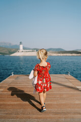 Little girl with a pink soft toy walks along the pier looking at the lighthouse. Back view