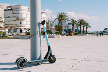 Rental blue electric scooter is fastened to a pole on an embankment with green palm trees
