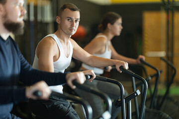 Fototapeta premium Concentrated athletic young adult man intensely working out on stationary air bicycle in fitness gym. Healthy active lifestyle concept