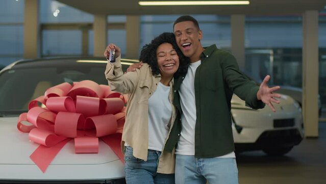 Happy smiling couple African American family woman man new car owners buyers showing keys at camera buying automobile with big bow at dealership store excited joyful male female buy luxury auto credit