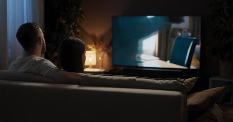 From behind young woman and man sit snuggled up together on couch watching romantic movie occasionally steal glanceing at each other hold hands hugging feeling content and relaxed. - Powered by Adobe