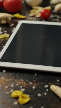 Camera panning right over farfalle pasta and fresh vegetables placed on table near contemporary white tablet PC. Vertical shot.