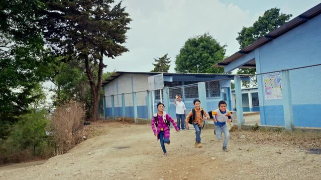 A group of happy children run in front of the school and the teacher says goodbye to them with a cordial greeting.