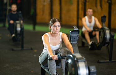 Determined young girl training with rowing machine in sports hall during crossfit workout