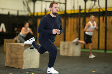Concentrated young bearded man in sport wear performing Bulgarian split squats on wooden plyometric...