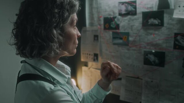 Medium close-up shot of mature Caucasian woman wearing shoulder holster standing in investigation bureau office, examining pin board with photos of suspects, analyzing murder and contemplating motive