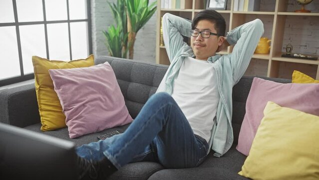 Young asian man relaxing on a couch in a modern living room, with colorful pillows and a relaxed posture.