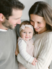 Fototapeta premium A happy family portrait with parents smiling at their baby, who is wearing a pink headband and looking at the camera. 